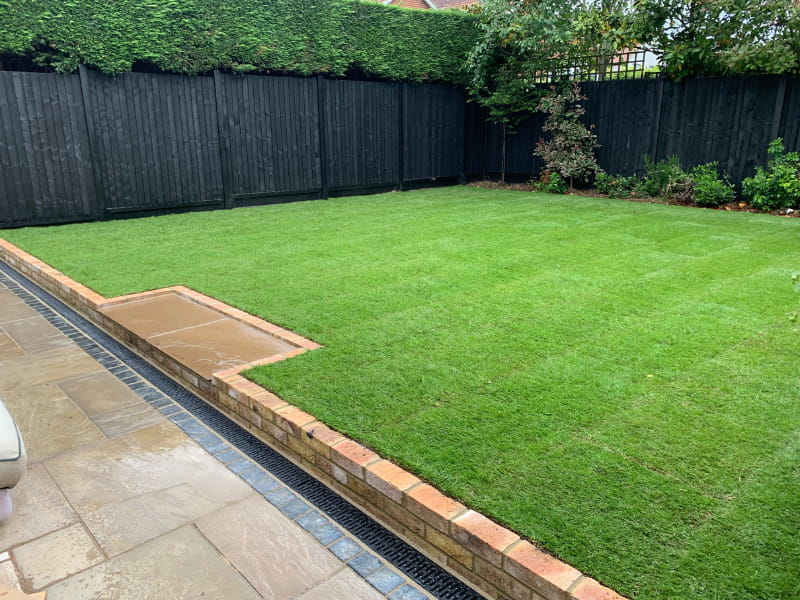 Large green lawn with brick edging beside stone patio and black wooden fence